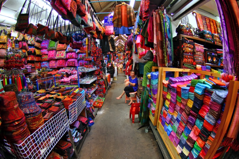 A vibrant market scene in Phnom Penh showcasing colorful textiles and handmade goods, with a vendor sitting in the aisle surrounded by an array of decorative fabrics and accessories.