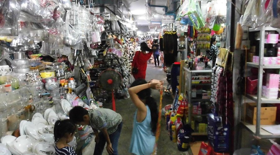 A bustling market aisle filled with various household items, including kitchenware, towels, and beauty products. Two children are seen interacting, while shoppers explore the extensive selection of goods.