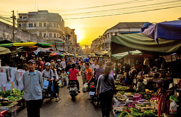 Vibrant night market scene in Phnom Penh with busy vendors, fresh produce, and colorful stalls under a sunset sky.