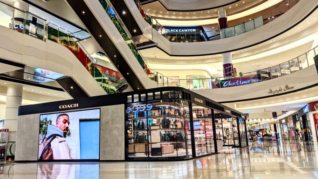 Interior view of a modern shopping mall featuring a Coach store with a large display screen, showcasing an advertisement. The mall has multiple levels with escalators visible and various shops in the background.