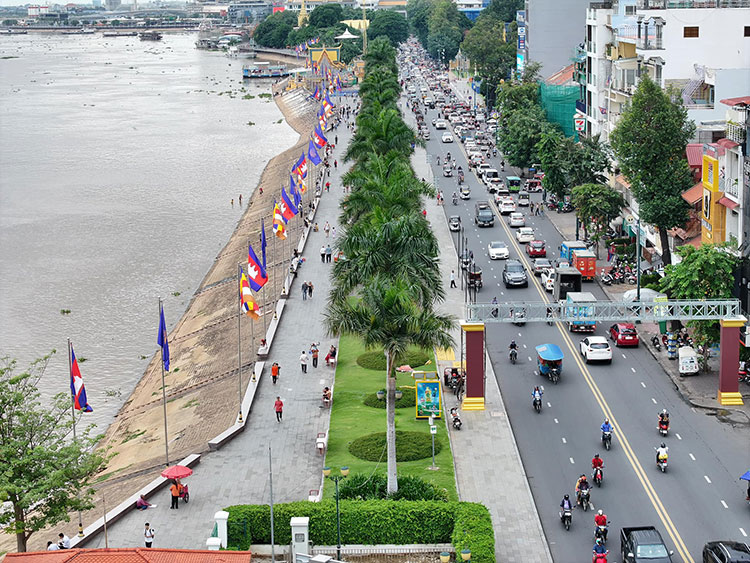 Aerial view of Riverside (Sisowath Quay) in Phnom Penh, featuring palm trees lining a paved promenade alongside the river, with colorful flags and a busy street filled with vehicles and pedestrians.