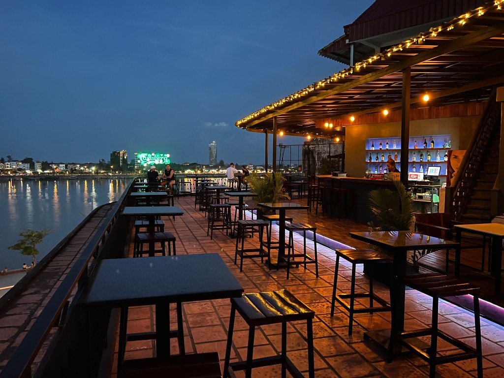Rooftop bar in Phnom Penh at night, featuring empty tables and a view of the river and city skyline.