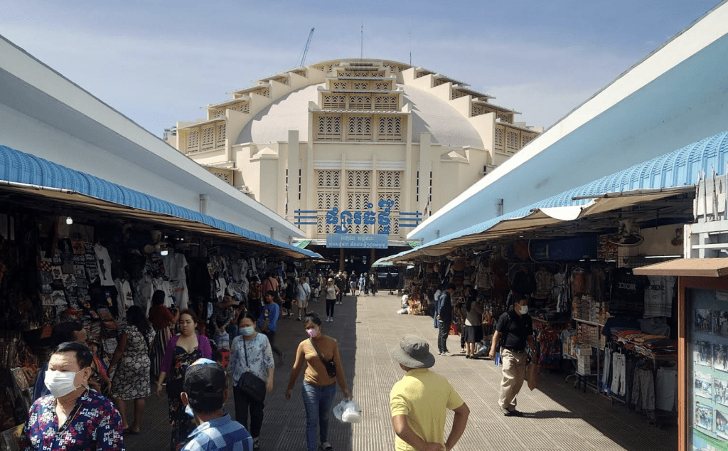 A bustling market scene in Phnom Penh, showcasing the vibrant Central Market filled with colorful stalls and shoppers navigating between rows of goods under a blue sky.