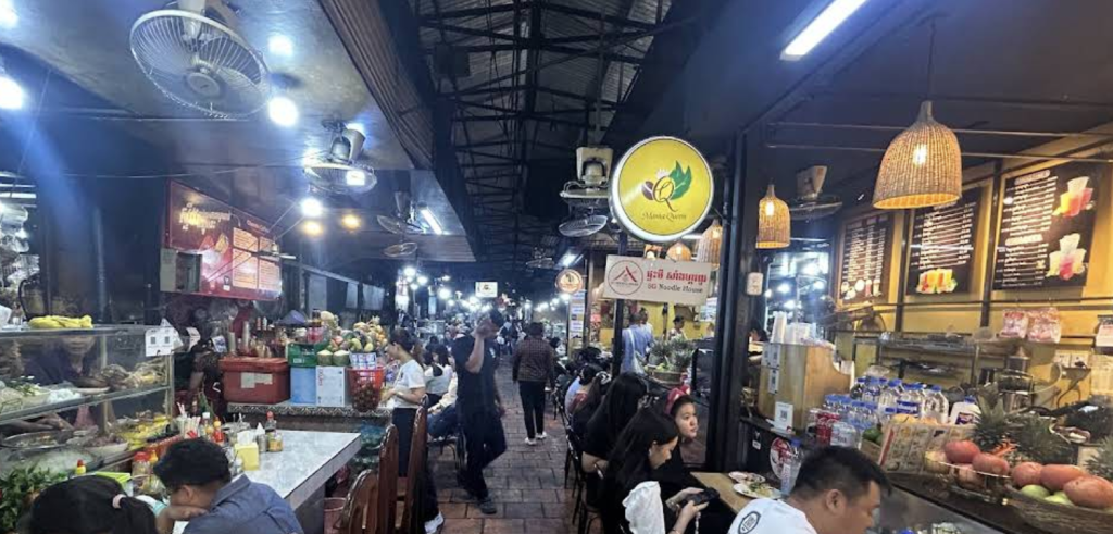 A bustling night market in Phnom Penh, featuring food stalls with various colorful dishes, customers enjoying their meals, and bright lights illuminating the scene.