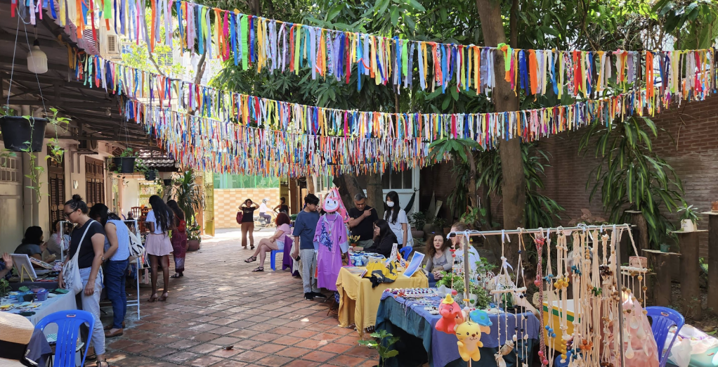 Artisan market scene in Phnom Penh, Cambodia, featuring colorful ribbon decorations above vendor tables, where people browse handmade crafts and local products.
