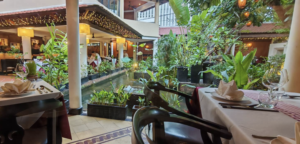View of an elegant dining area surrounded by lush greenery and a water feature, with tables set for a meal in a well-decorated restaurant.