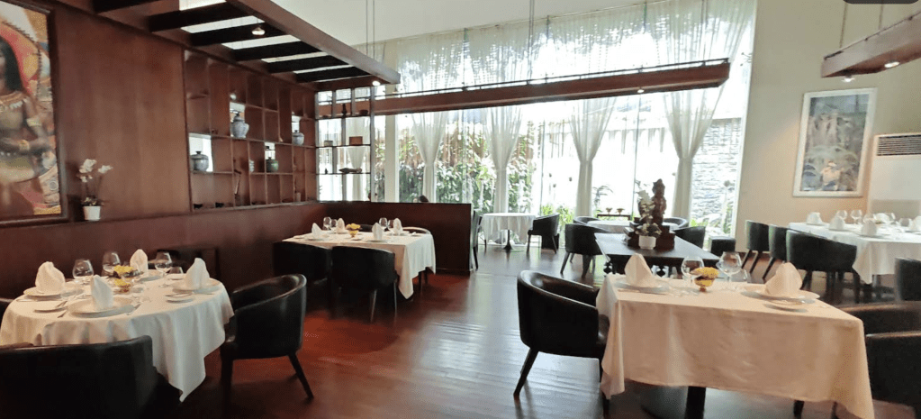 Elegant dining room with polished wooden floors, neatly set tables, and large windows allowing natural light, featuring a decorative wall shelf and greenery visible outside.
