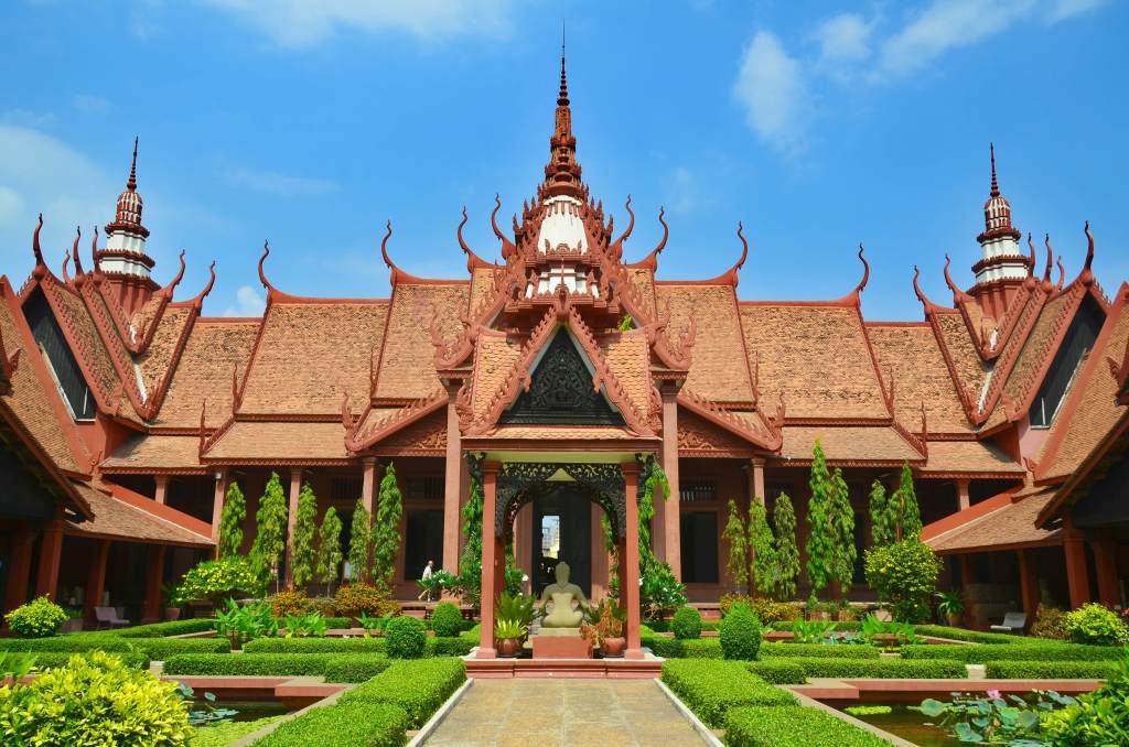 A vibrant view of the National Museum of Cambodia, showcasing its traditional Khmer architecture surrounded by lush greenery and manicured gardens, under a clear blue sky.