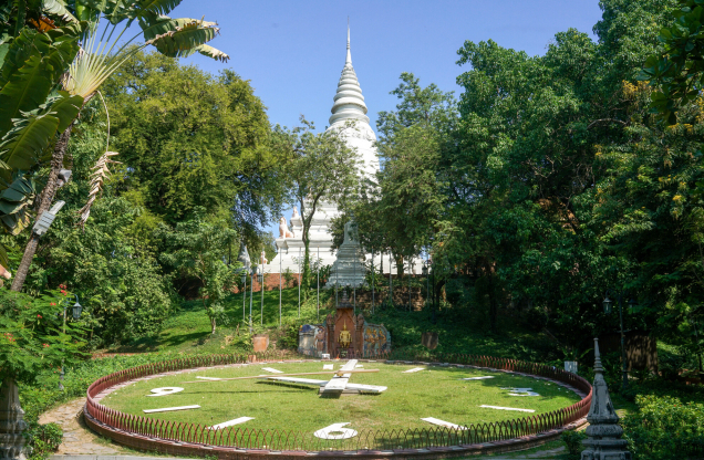 A scenic view of a clock face made of grass and stone, surrounded by greenery and featuring a white stupa in the background.