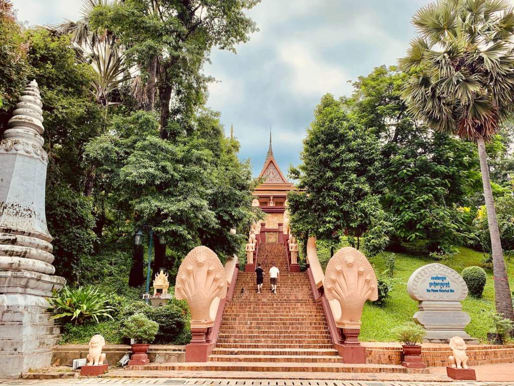 A scenic view of the steps leading up to Wat Phnom, surrounded by lush greenery and palm trees, with decorative lion statues on either side.