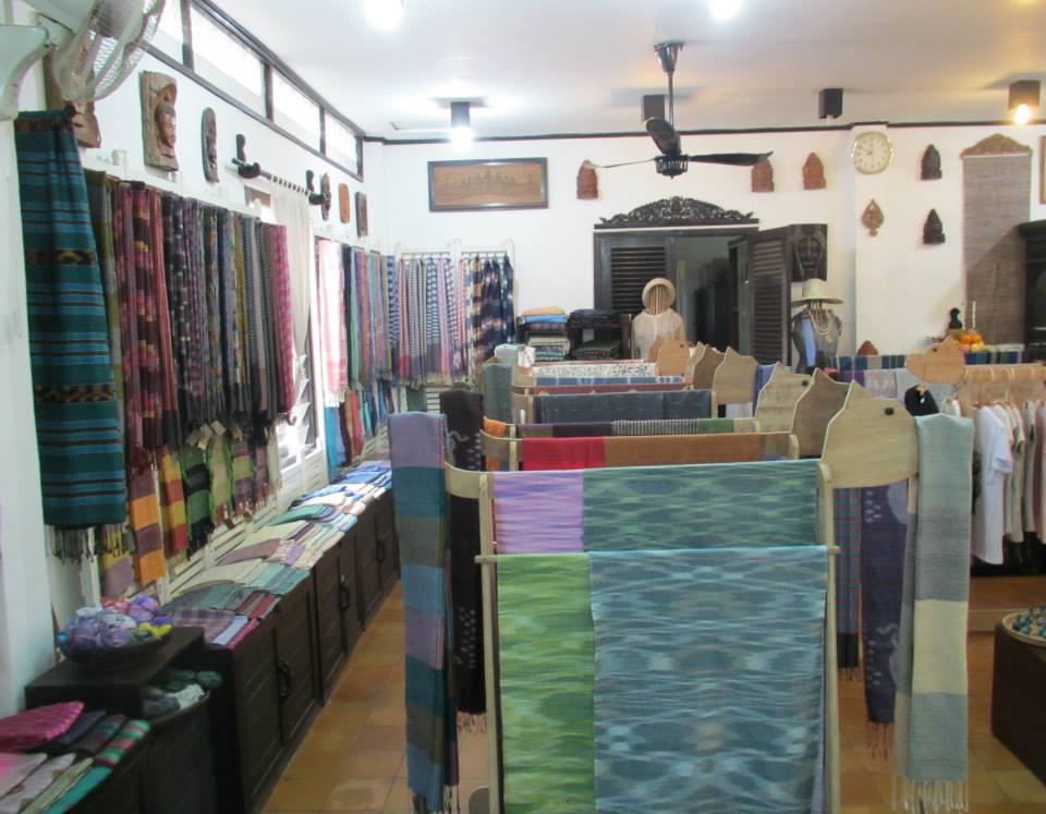 Inside a craft shop showcasing an array of colorful handwoven scarves and textiles, displayed on racks and walls, with wooden furniture and decorative artifacts in the background.