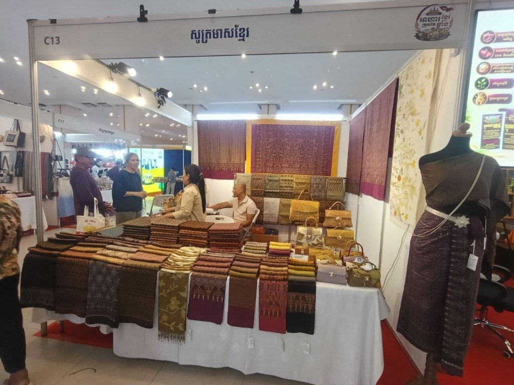 A Khmer silk and textile stall at the Khmer Food Must Be Strong exhibition, displaying traditional sampots, scarves and woven fabrics with a mannequin dressed in a Khmer outfit