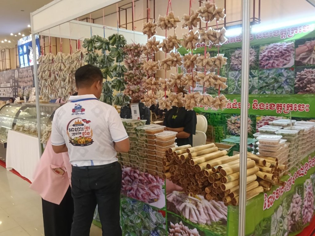 A Khmer vendor standing beside a stall filled with traditional dried snacks, bamboo shoots, sticky rice cones and local food products at the Khmer Food Must Be Strong exhibition 2026
