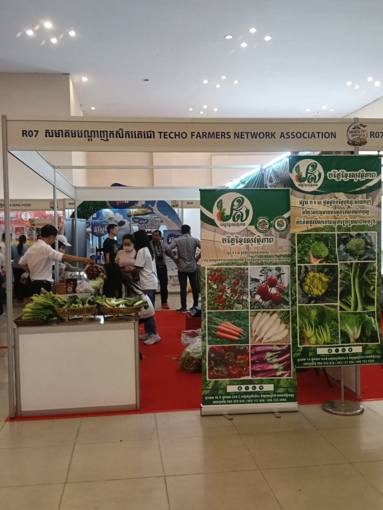 A fresh vegetable stall at the Khmer Food Must Be Strong exhibition, with a large display board showing locally grown Cambodian vegetables and visitors browsing the produce