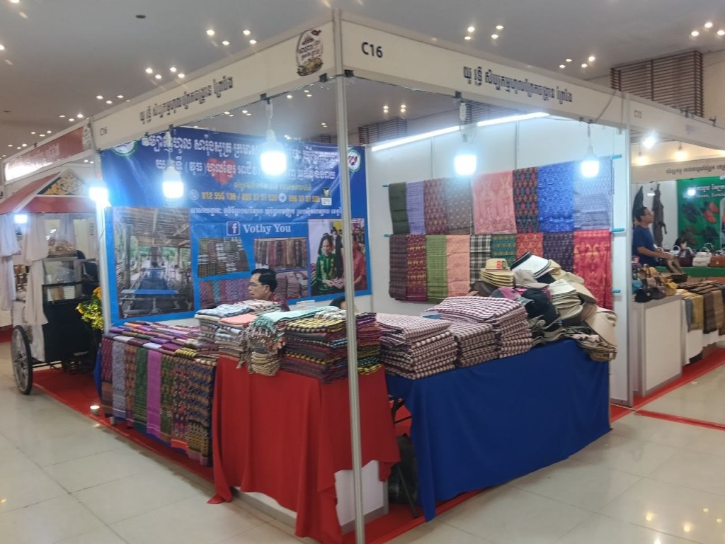 The Vothy You textile stall at the Khmer Food Must Be Strong exhibition, displaying colourful Cambodian woven fabrics, krama scarves and traditional patterned cloth at Koh Pich Theater 2026