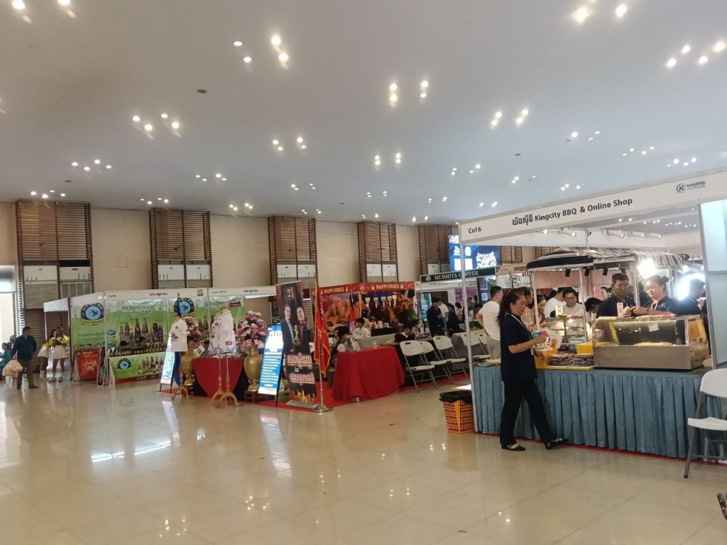 A wide-angle view of the Khmer Food Must Be Strong exhibition hall at Koh Pich Theater, showing dozens of Cambodian food and product stalls with visitors browsing under the grand lit ceiling