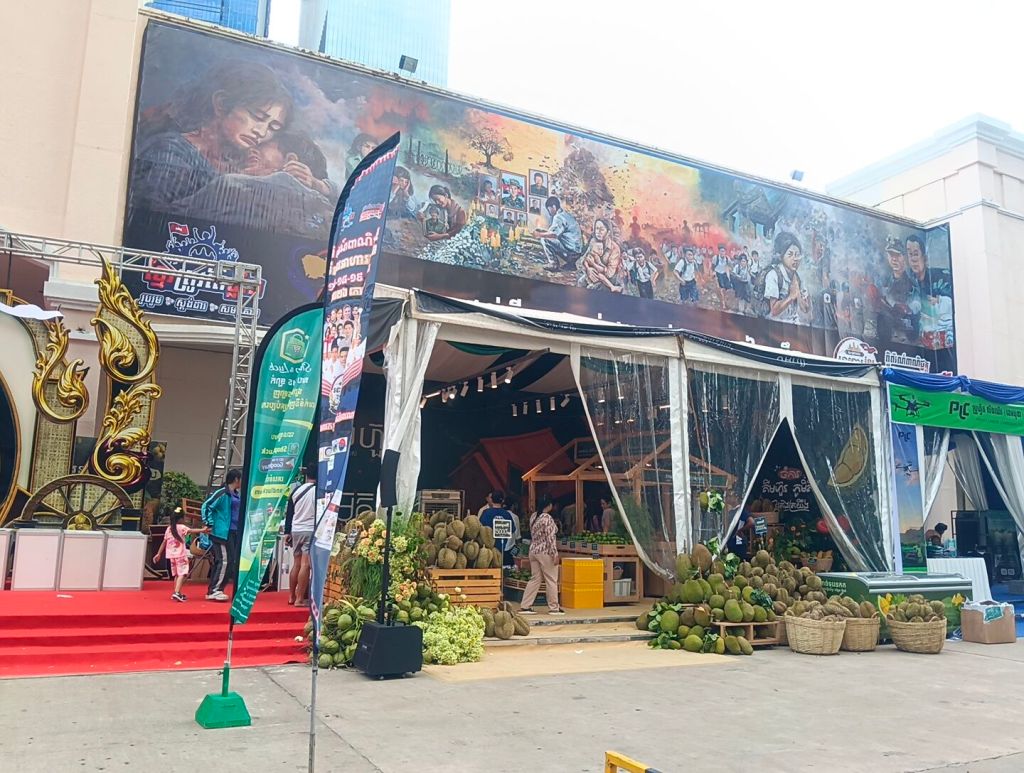 The outdoor entrance of the Khmer Food Must Be Strong exhibition at Koh Pich Theater, with fresh coconuts and durians displayed at the front and a large Khmer mural on the building