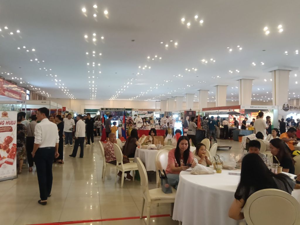 Visitors dining and socialising at tables inside the Khmer Food Must Be Strong exhibition hall at Koh Pich Theater, with Cambodian food and product stalls filling the background