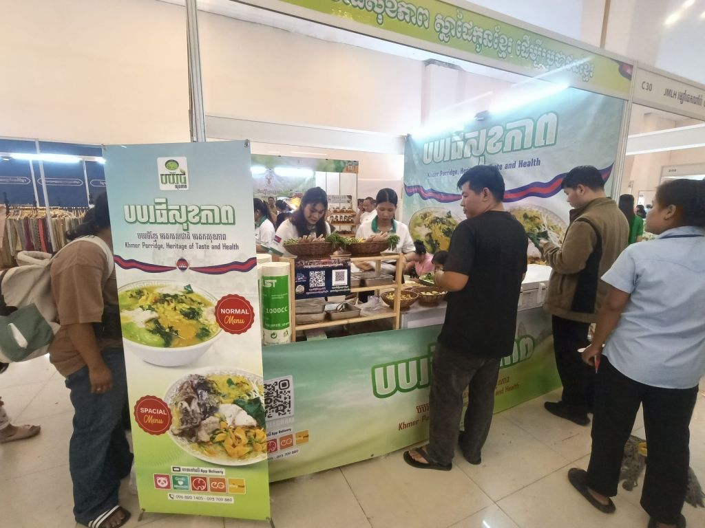 Visitors trying Borbor Sromal Khmer porridge at the Khmer Food Must Be Strong exhibition, with a branded green stall and menu banners showing traditional Cambodian rice porridge dishes at Koh Pich Theater 2026