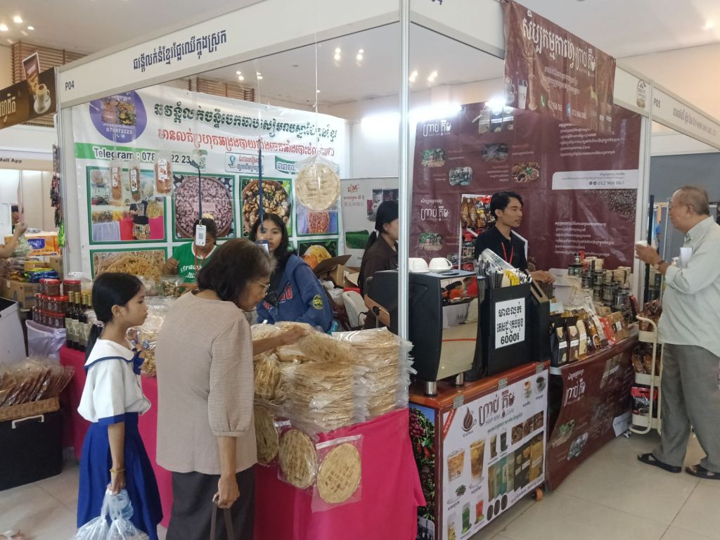 Visitors browsing Cambodian food and product stalls at the Khmer Food Must Be Strong exhibition at Koh Pich Theater, with a vendor serving an elderly customer at a local food products booth